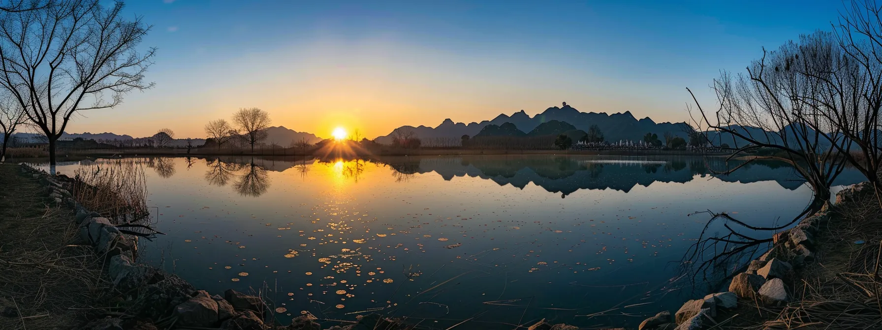 a vivid sunset casts warm golden hues over a serene lake, reflecting the towering silhouettes of surrounding mountains under a clear blue sky.