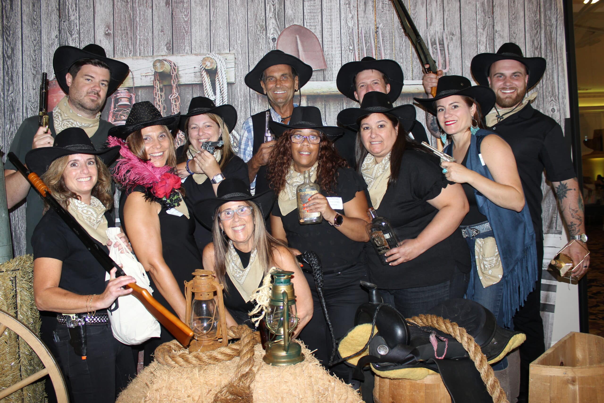 A group of people dressed in cowboy hats and bandanas pose together in a western-themed setting with props like lanterns, ropes, and a saddle.