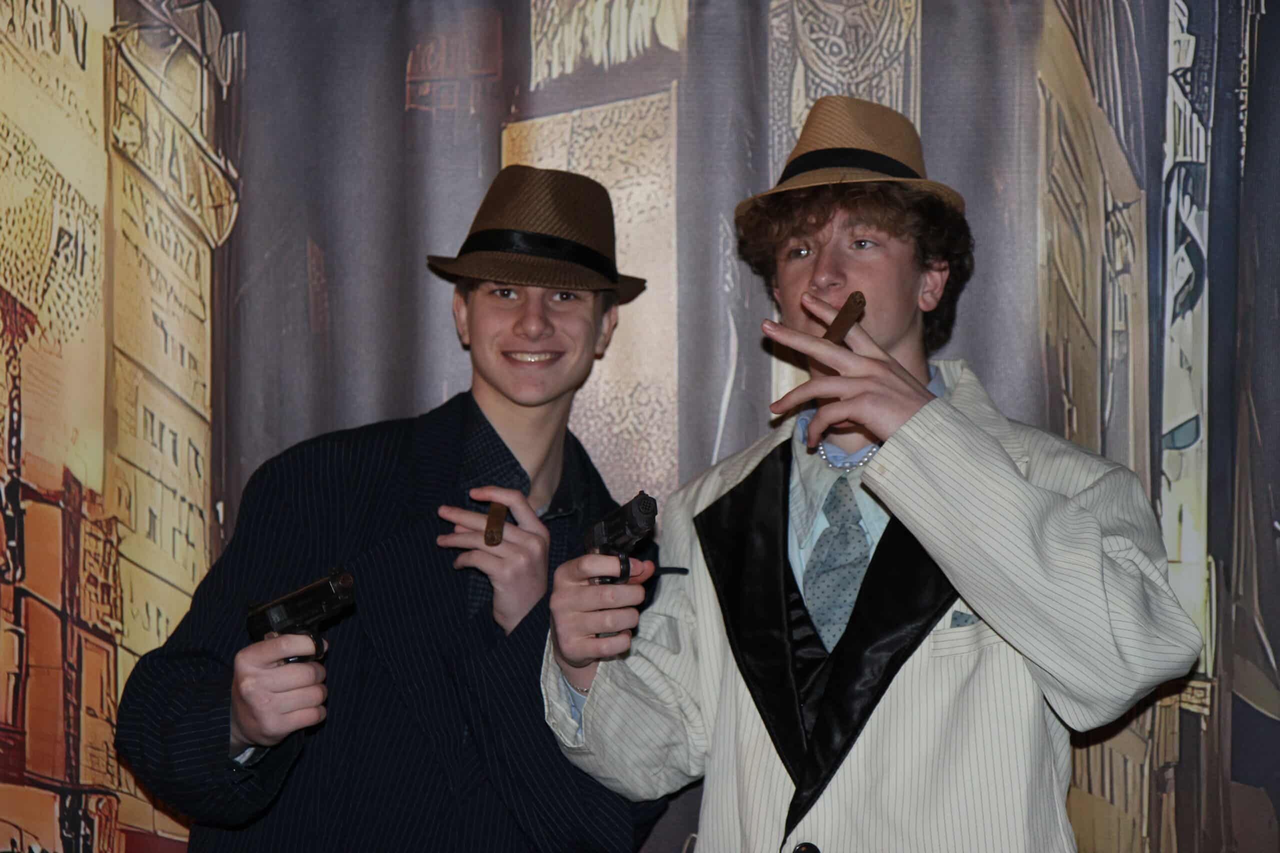 Two young men dressed in suits and fedoras pose with toy guns and cigars, standing in front of a city backdrop, evoking a gangster or 1920s theme.