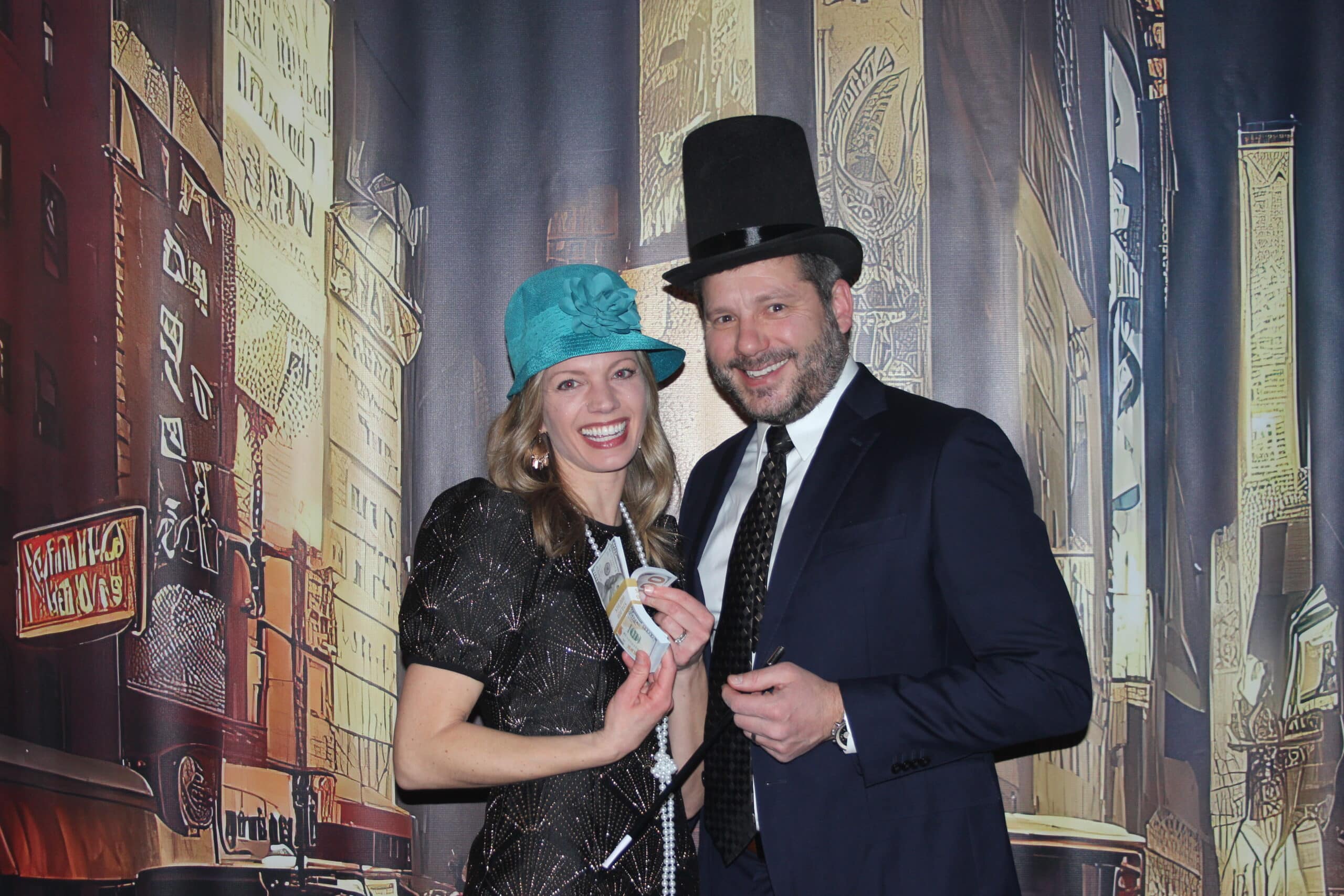 A smiling woman in a blue hat and a man in a black top hat and suit pose together in front of a vintage cityscape backdrop, holding money and a cigarette holder, dressed in a 1920s-themed style.
