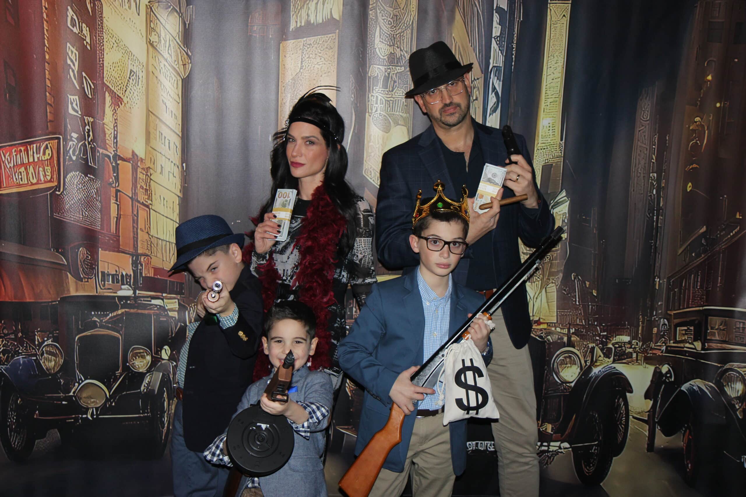A family in 1920s gangster costumes poses with toy guns, crowns, and fake money against a vintage city backdrop with classic cars.