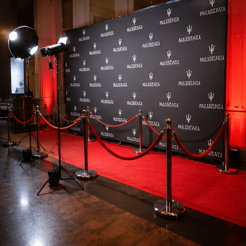 A red carpet event setup with stanchions, red ropes, bright lights, and a backdrop featuring Maserati logos in a grand, warmly-lit venue.