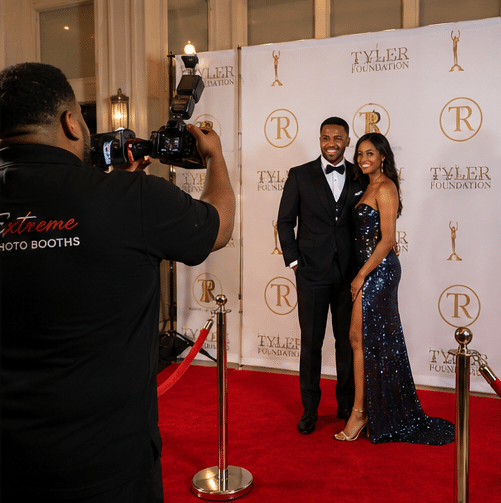 A photographer takes a picture of a smiling couple dressed formally on a red carpet at a gala event, with a Tyler Foundation backdrop behind them.