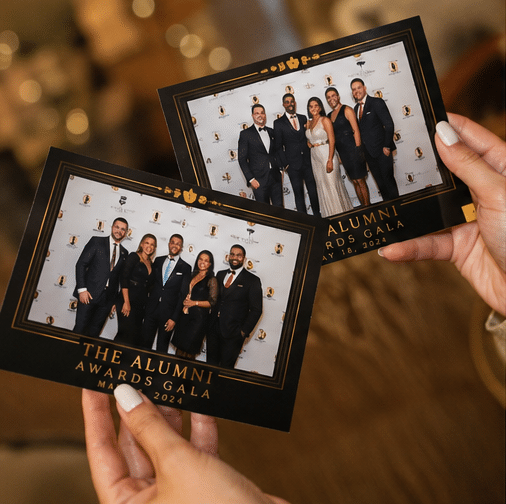 Two hands hold photo prints from the Alumni Awards Gala, showing groups of people in formal attire standing together and smiling for the camera.