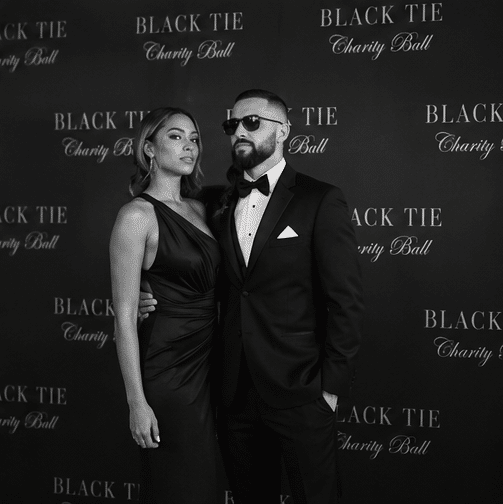 A woman in a black dress and a man in a black tuxedo and sunglasses pose together at a Black Tie Charity Ball event backdrop.